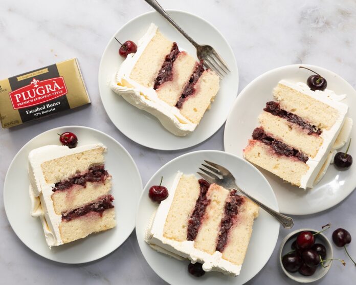 Slices of cherry layer cake on white plates with whipped frosting and fresh cherries nearby; butter box in corner for context of ingredients? (informative food image)