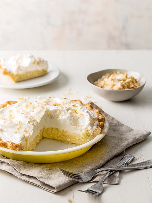 Coconut Cream Pie in pie dish with slice of pie on white plate, bowl of shaved coconut, and forks