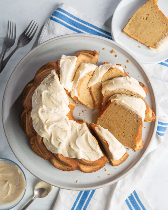 Irish Cream Pound Cake on white platter with forks, a spoon, a bowl of cream, a slice of pound cake on a white plate, and a blue striped dish towel