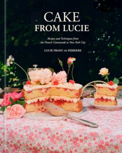 Pink, two-layer cake with pink frosting and fresh flowers on a pink floral tablecloth outdoors, with the text 'CAKE FROM LUCIE' in the dark background.