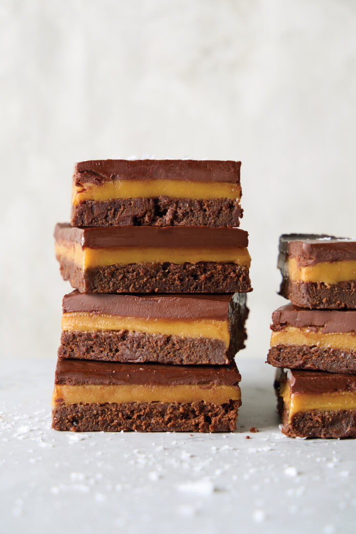Stack of four chocolate brownie bars with caramel filling and a glossy chocolate top on a light surface.