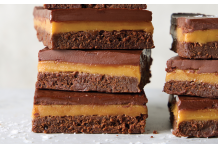 Millionaire Brownie Bars Stack of four chocolate brownie bars with caramel filling and a glossy chocolate top on a light surface.