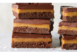 Millionaire Brownie Bars Stack of four chocolate brownie bars with caramel filling and a glossy chocolate top on a light surface.
