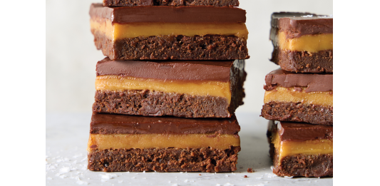 Millionaire Brownie Bars Stack of four chocolate brownie bars with caramel filling and a glossy chocolate top on a light surface.