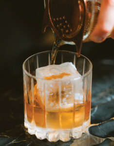 Person pours amber liquid from a copper jigger into a glass with ice, close-up.