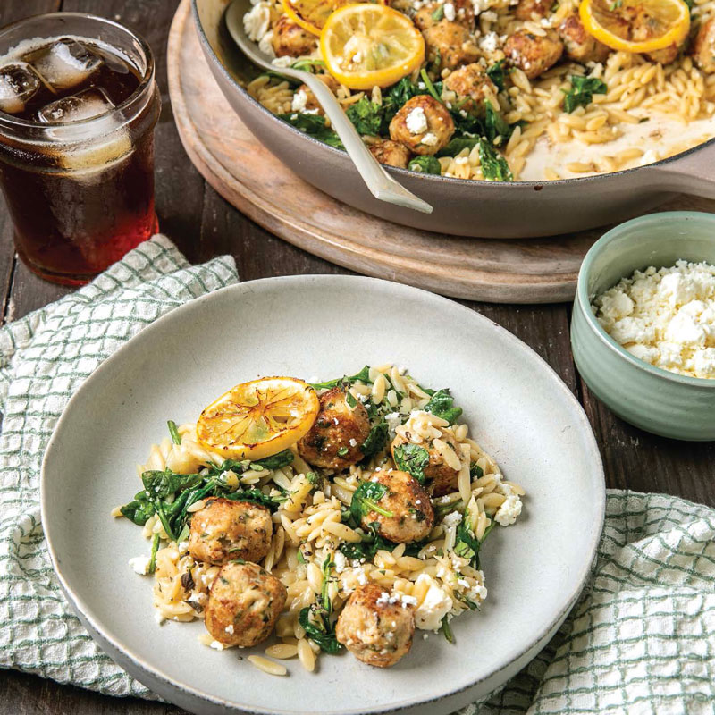 Plate of orzo with meatballs, spinach, and crumbled cheese topped with a lemon slice on a light plate. A skillet of the same dish and a glass of iced tea are in the background.