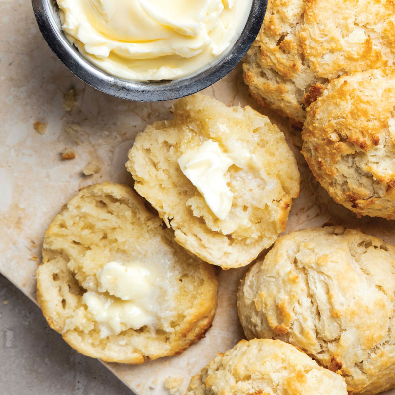 Buttery biscuits or scones with pats of melted butter on a wooden board and a small butter cup in the corner.