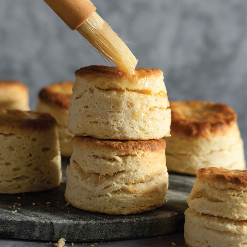 Stack of light, fluffy scones on a dark board with melted butter being brushed onto the top