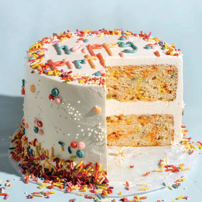 Two-layer white-frosted birthday cake with rainbow sprinkles on the rim; a slice is cut to reveal yellow-orange sponge with colorful bits inside, set against a light blue background.
