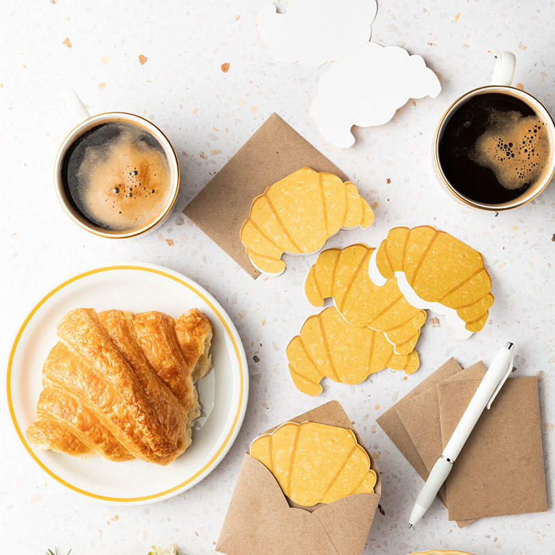 Top-down breakfast scene with two cups of coffee, a croissant on a plate, and yellow crescent-shaped crackers on brown wrappers.