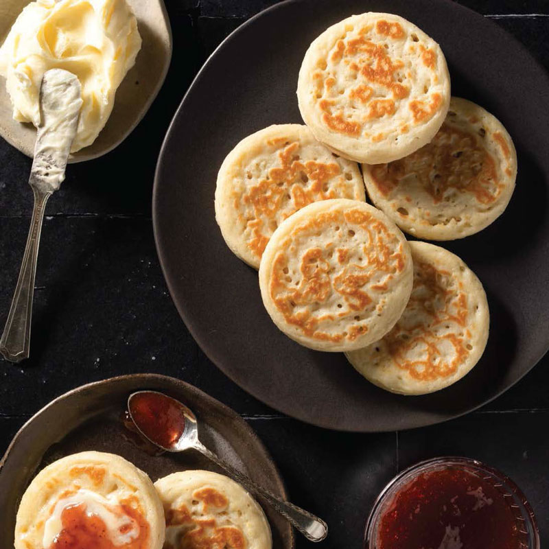 Stack of golden-brown English muffins on a dark plate, with butter on the side and a jar of jam nearby for breakfast.