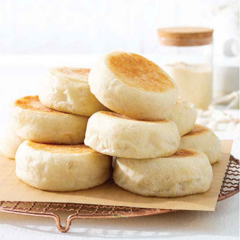 Stack of lightly browned English muffins piled on parchment on a cooling rack, with a jar in the soft background.