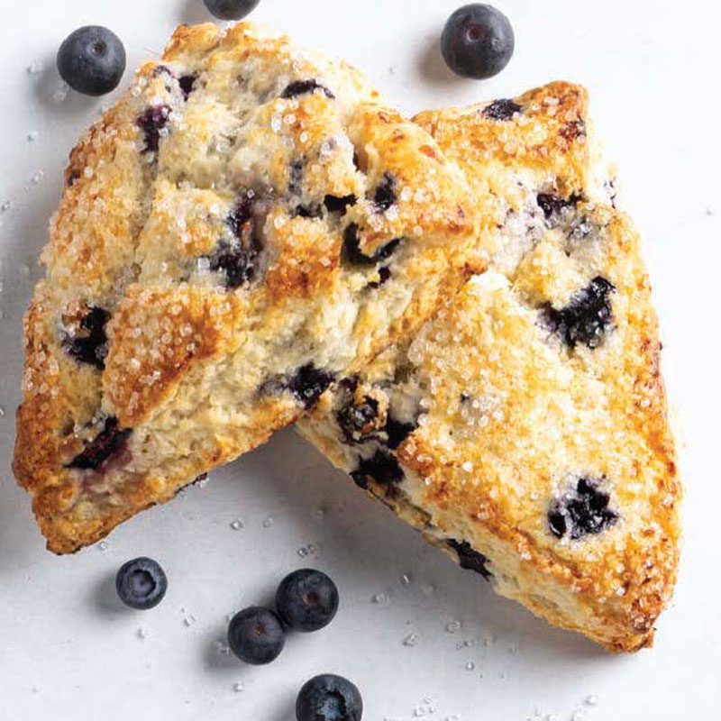 Two blueberry scones broken apart on a white surface, flecked with sugar crystals and whole blueberries nearby.