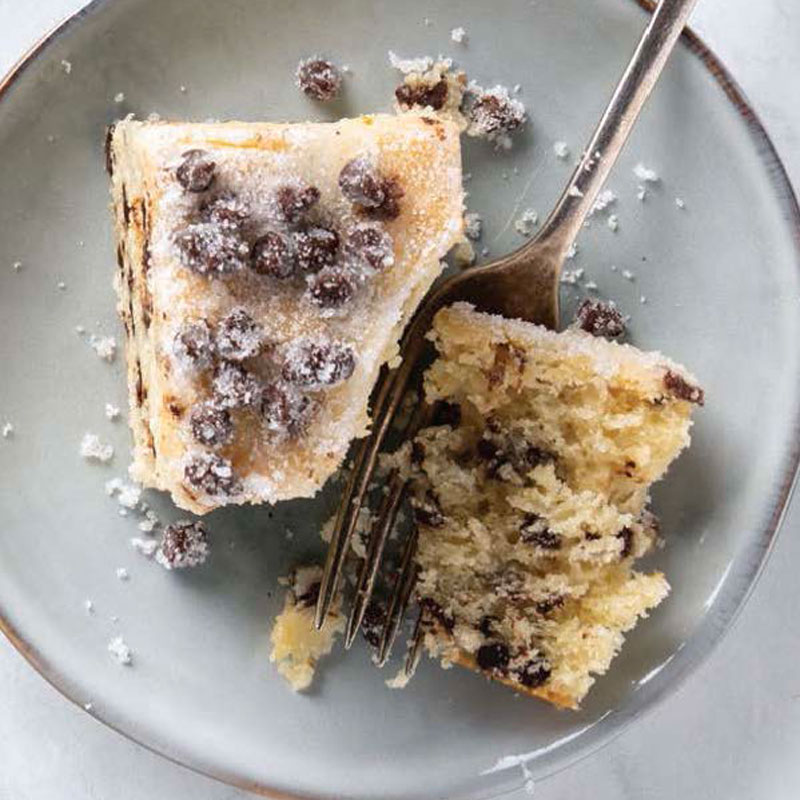 Two slices of vanilla cake with chocolate chips on a pale blue plate, a fork resting beside them and a dusting of sugar crystals visible on top.