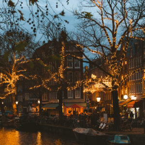 Amsterdam Evening city canal scene with leafless trees wrapped in warm string lights and glowing reflections on the water along brick buildings.",
