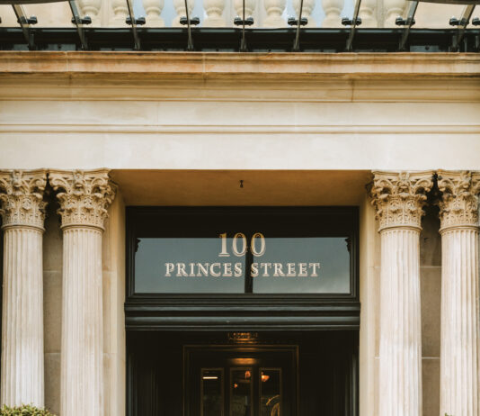 Edinburgh’s Cozy and Exclusive New Address Doorman in a dark suit and hat stands on stone steps in front of a grand doorway with decorative columns and a 100 Princes Street sign above.