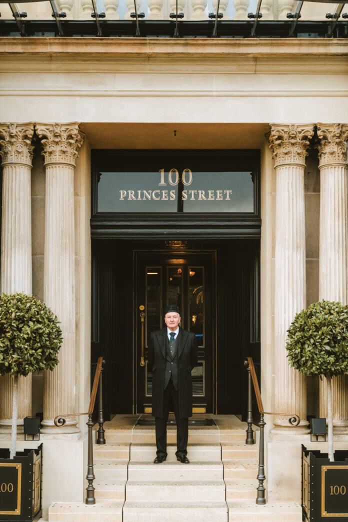 Doorman in a dark suit and hat stands on stone steps in front of a grand doorway with decorative columns and a 100 Princes Street sign above.