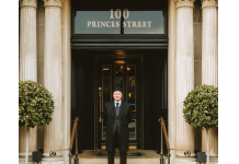 Edinburgh’s Cozy and Exclusive New Address Doorman in a dark suit and hat stands on stone steps in front of a grand doorway with decorative columns and a 100 Princes Street sign above.