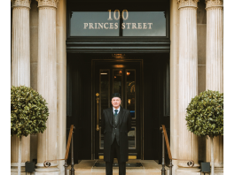 Edinburgh’s Cozy and Exclusive New Address Doorman in a dark suit and hat stands on stone steps in front of a grand doorway with decorative columns and a 100 Princes Street sign above.