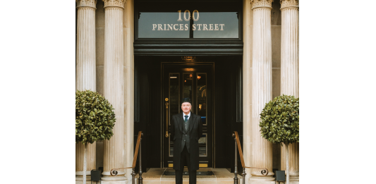 Edinburgh’s Cozy and Exclusive New Address Doorman in a dark suit and hat stands on stone steps in front of a grand doorway with decorative columns and a 100 Princes Street sign above.