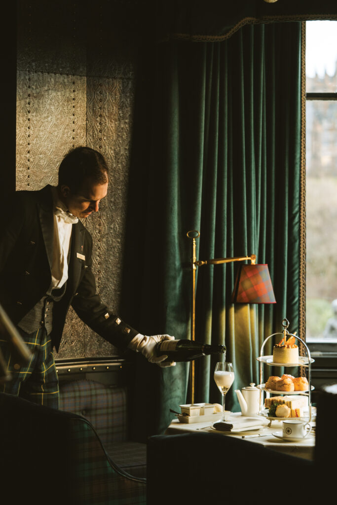 Man in a tuxedo pours champagne at a table with desserts and tea setup in a dimly lit, elegant room.