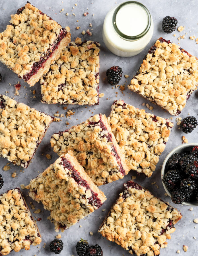 Crumbled blackberry bars scattered on a gray surface with a small pitcher of milk and fresh blackberries nearby.