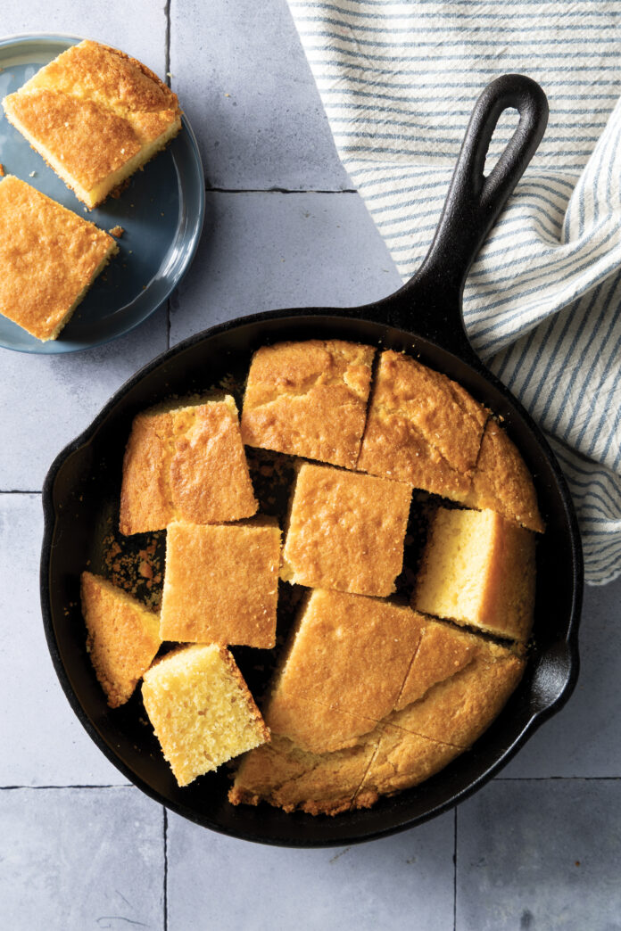Cast iron skillet filled with golden cornbread squares on a light blue surface, with a striped cloth nearby.