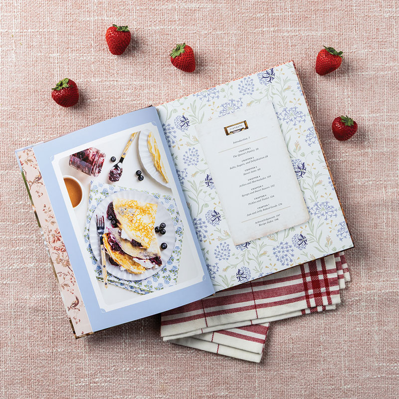 Open cookbook on pink fabric, plate of pancakes with berries, floral page, and napkins nearby.