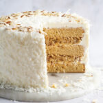Two-layer coconut cake with white frosting and toasted almond shavings, sliced to reveal creamy filling between sponge layers on a marble surface.