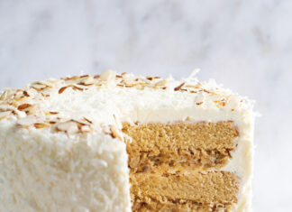 Creamy Coconut-Almond Layer Cake Two-layer coconut cake with white frosting and toasted almond shavings, sliced to reveal creamy filling between sponge layers on a marble surface.
