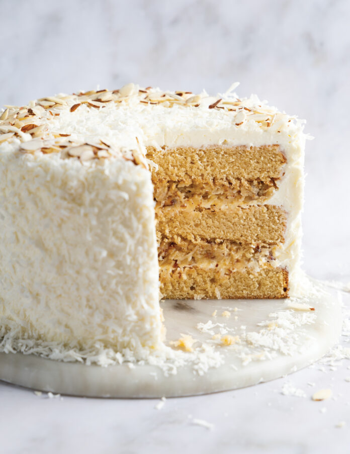 Two-layer coconut cake with white frosting and toasted almond shavings, sliced to reveal creamy filling between sponge layers on a marble surface.