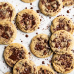 Several chocolate chip cookies arranged on white parchment on a light surface, with chocolate crumbs scattered around.