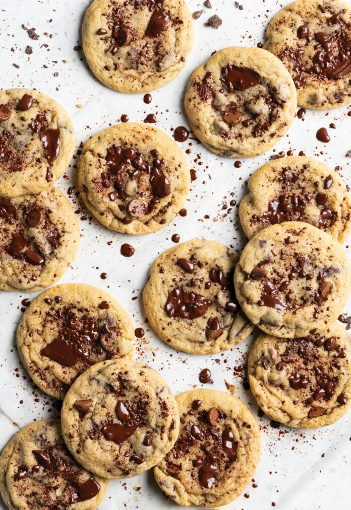 Several chocolate chip cookies arranged on white parchment on a light surface, with chocolate crumbs scattered around.