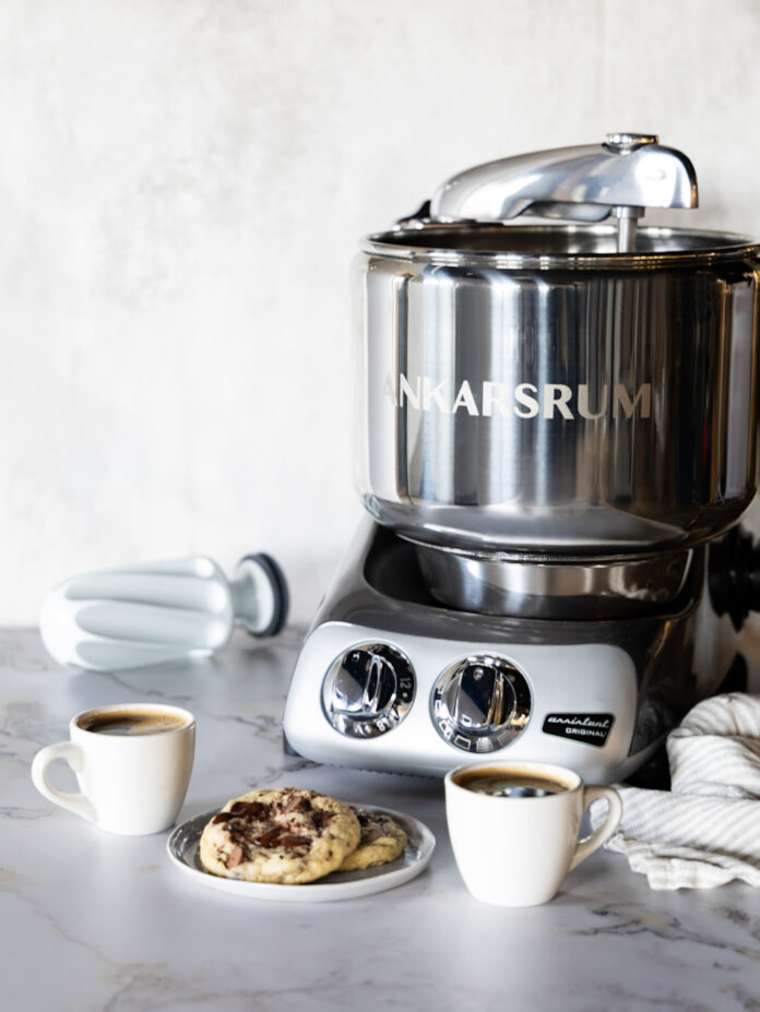 Stainless steel Ankarsrum stand mixer on a marble kitchen counter, with coffee cups and cookies nearby.