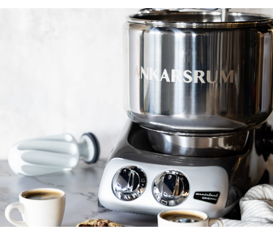 Ankarsrum Recipe Conversions Stainless steel Ankarsrum stand mixer on a marble kitchen counter, with coffee cups and cookies nearby.