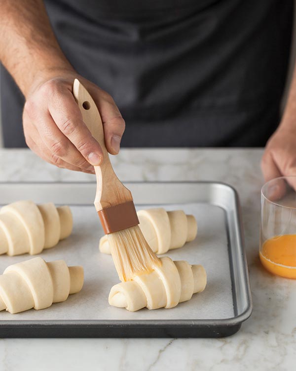 Egg wash being brushed onto shaped croissants