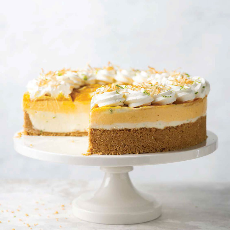 Cheesecake with crumb crust on a white pedestal, topped with piped cream and citrus zest and a lemon layer visible inside.