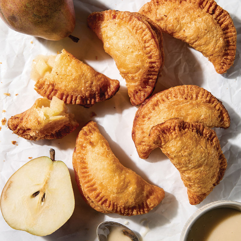 Golden baked empanadas on parchment with a pear half and a small bowl of sauce nearby.
