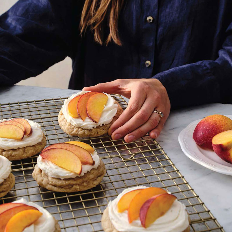 Person decorating peach-trost cream cookies on a wire rack, with a plate of peaches nearby.