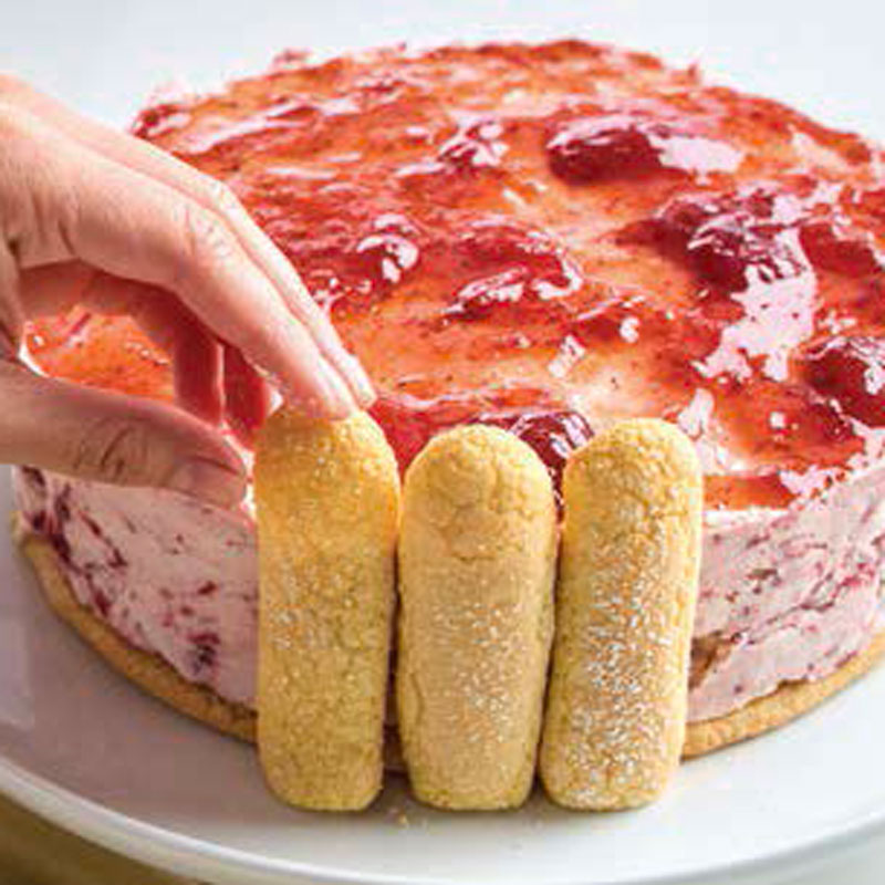 Hand placing ladyfingers around a cherry-topped cheesecake on a plate.
