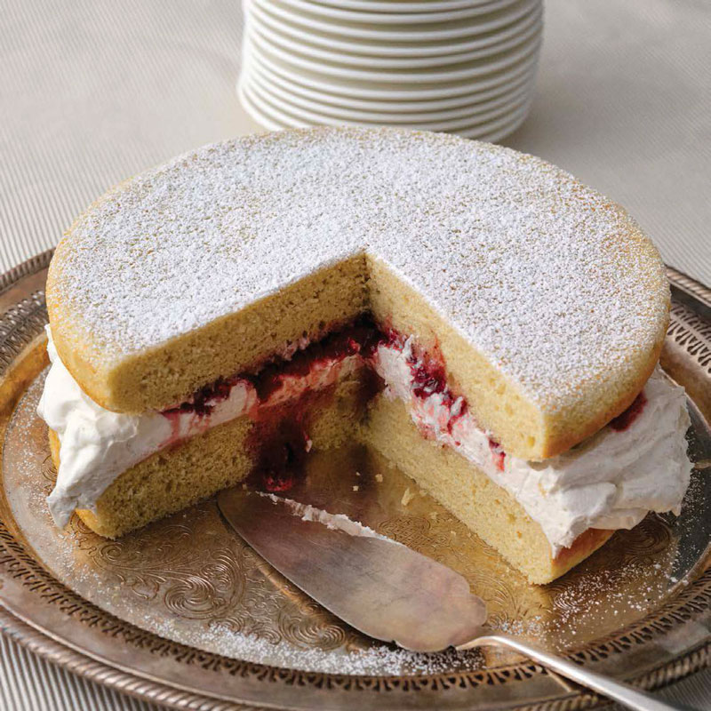 Vanilla sponge cake filled with whipped cream and berry jam, lightly dusted with powdered sugar on a decorative plate, with a slice removed.
