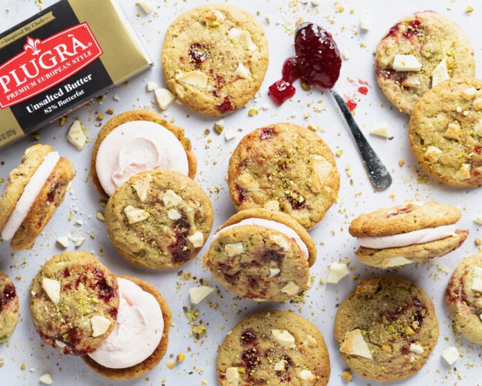 Close-up of assorted cookies with jam centers, white chocolate chunks, and pistachio crumble next to a butter box.