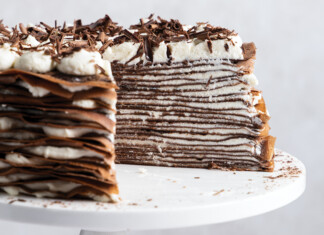 Chocolate Crêpe Cake with Mascarpone Whipped Cream Slice of multi-layer chocolate cake with cream filling and chocolate shavings on a white cake stand
