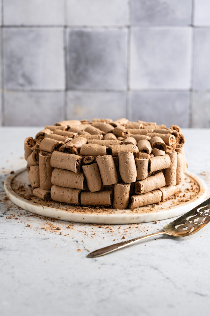 Chocolate wafer-roll cake arranged in a circle on a white plate, with crumbs scattered nearby on a light countertop