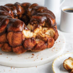 Glossy chocolate-glazed monkey bread bundt on a white plate, with two coffee cups in the background.