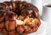 Monkey Bread Glossy chocolate-glazed monkey bread bundt on a white plate, with two coffee cups in the background.