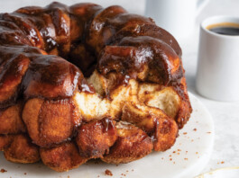 Monkey Bread Glossy chocolate-glazed monkey bread bundt on a white plate, with two coffee cups in the background.