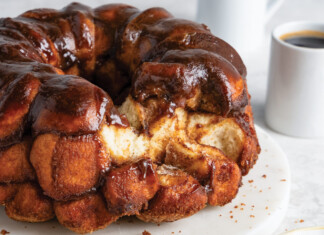 Monkey Bread Glossy chocolate-glazed monkey bread bundt on a white plate, with two coffee cups in the background.