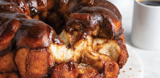 Monkey Bread Glossy chocolate-glazed monkey bread bundt on a white plate, with two coffee cups in the background.