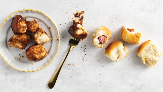 Top-down view of a white plate with golden fried chicken bites, a fork, and several bread rolls on a light textured surface.